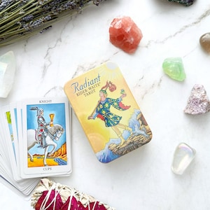 May include: A Rider-Waite Tarot deck in a metal tin with a colorful illustration of a figure walking on a mountain. The deck is next to a stack of tarot cards, crystals, and a bundle of sage.
