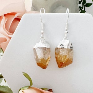 May include: A pair of silver earrings with raw citrine crystal drops. The crystals are a light orange color and have a natural, rough texture.
