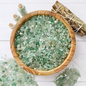 May include: A wooden bowl filled with green and clear tumbled stones. The bowl is on a white surface with a bundle of dried herbs and two small glass bottles.