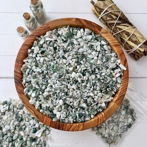 May include: A wooden bowl filled with green and white tumbled stones. The stones are scattered around the bowl on a white surface.