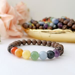 May include: A brown wooden bracelet with a copper clasp and seven colorful gemstone beads. The beads are arranged in a rainbow pattern, starting with red and ending with purple.