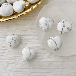 May include: Five white heart-shaped stones with gray veining. The stones are arranged on a white surface and a gold plate.