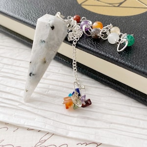 May include: A white crystal pendulum with a silver chain and a rainbow of chakra stones attached. The pendulum is on a white surface with a black notebook in the background.