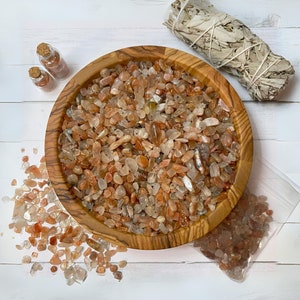 May include: A wooden bowl filled with orange and clear sunstone chips. The bowl is on a white wooden surface. There is a small bag of sunstone chips and a bundle of dried sage next to the bowl.