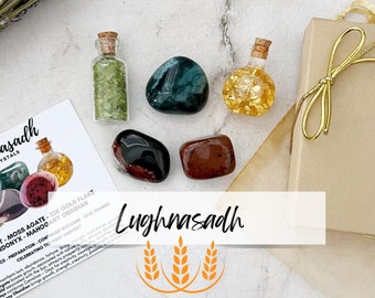 May include: A collection of crystals including a small glass bottle filled with green peridot chips, a dark green moss agate, a small glass bottle with gold flakes, a black sardonyx, and a mahogany obsidian stone, alongside a Lughnasadh card.