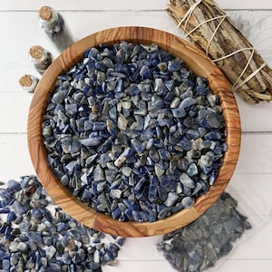 May include: A wooden bowl filled with blue and white tumbled stones. The bowl is on a white surface with a small bag of stones and two glass bottles.