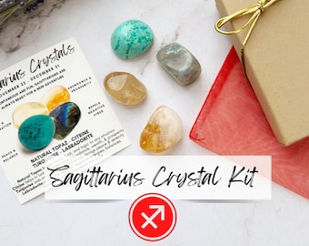 May include: A Sagittarius crystal kit with a red Sagittarius symbol, a small brown gift box, and five crystals: a turquoise, a clear crystal, a yellow crystal, a brown crystal, and a gray crystal. The crystals are arranged on a white surface with a lavender sprig in the background. The text on the card reads "Sagittarius Crystals November 22 - December 21 Spontaneous and fun, Sagittarians are always ready for a new adventure Abundance & Prosperity Repels Negative Vibes Natural Topaz - Citrine Turquoise - Labradorite Natural Topaz brings abundance, wealth, & prosperity Citrine helps in luck, and logic to any situation Turquoise aids or repels negative energies, balances. Labradorite"