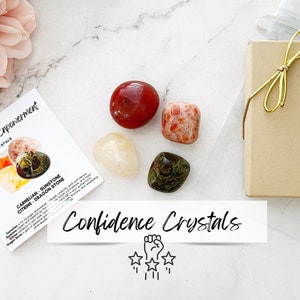 May include: A set of four crystals in a small box. The crystals are carnelian, sunstone, citrine, and dragon stone. The box is brown with a gold ribbon. The crystals are arranged in a circle with a white card in the center that says "Confidence Crystals".