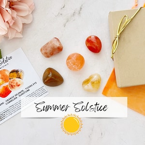 May include: A set of six polished crystals in shades of orange, yellow, and red. The crystals are arranged on a white surface with a small card that reads "Summer Solstice" and a small gift box with a gold ribbon. The crystals are labeled as Citrine, Orange Selenite, Tigers Eye, Carnelian, and Sunstone.
