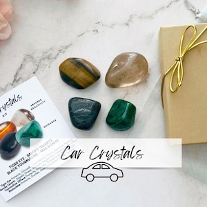 May include: A set of five crystals in a small gift box. The crystals are tiger eye, smoky quartz, black tourmaline, malachite, and clear quartz. The box is brown with a gold ribbon. The crystals are arranged in a circle on a white surface. The text "Car Crystals" is written in black on a white background.