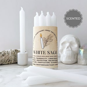 May include: A collection of white spell candles with a "White Sage" label, a white skull, and a bundle of sage. The label includes the text "The Stone Sanctuary Shop Established 2017" and "Scented".