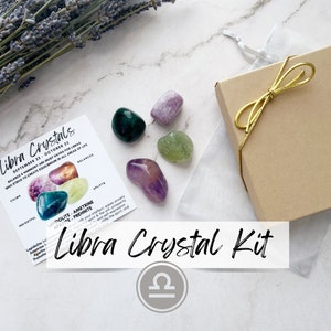 May include: A Libra Crystal Kit with five crystals in a small box. The crystals are amethyst, green aventurine, lepidolite, prehnite, and ametrine. The crystals are arranged on a white surface with a card that says "Libra Crystals" and lists the crystals included in the kit. The Libra symbol is in the lower right corner of the image.