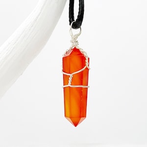 May include: A vibrant orange crystal pendant with a silver wire wrap, suspended from a black leather cord. The crystal is a long, hexagonal shape, with a pointed tip. The pendant is displayed against a white background.