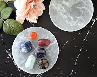 May include: A set of seven colorful crystals arranged on a white selenite charging plate. The crystals include a red carnelian, a blue sodalite, a red jasper, a purple amethyst, a green aventurine, a black obsidian, and a clear quartz.