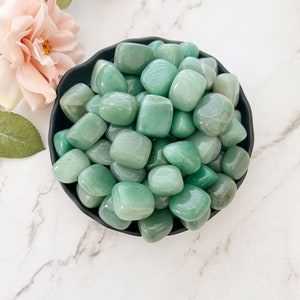 May include: A bowl of green tumbled stones. The stones are smooth and polished, and they are arranged in a random pattern. The bowl is black and has a glossy finish.