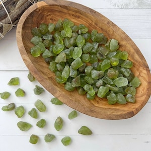 May include: A wooden bowl filled with raw, olive-green peridot gemstones. Some stones are scattered outside the bowl. The bowl has a natural wood grain and sits on a white surface. A bundle of dried sage is in the background.