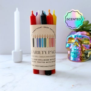 May include: A variety pack of scented spell candles in a brown paper sleeve. The candles are in various colors, including red, pink, black, orange, and yellow. The sleeve has text that reads "Variety Pack" and "Scented Spell Candles". A white candle is in a holder to the left.