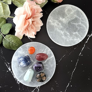 May include: A set of seven colorful crystals arranged on a white selenite charging plate. The crystals include a red carnelian, a blue sodalite, a red jasper, a purple amethyst, a green aventurine, a black obsidian, and a clear quartz.