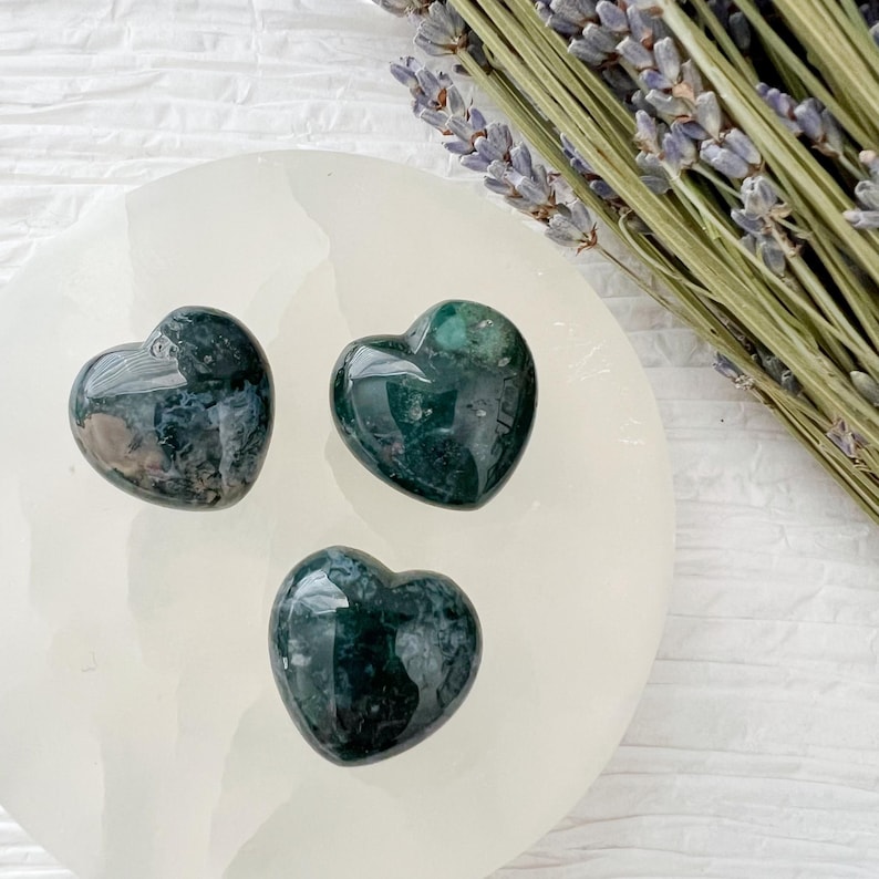 May include: Three heart-shaped stones with a dark green and white pattern are arranged on a white, circular surface. A bundle of dried lavender is placed to the side. The stones have a polished, glossy finish.