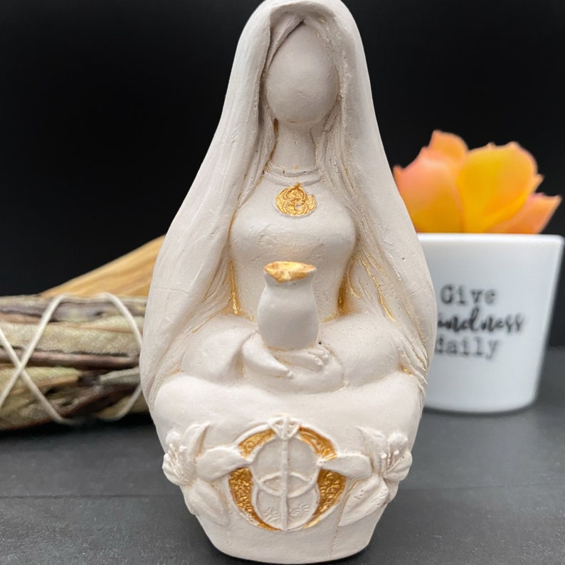 Puede incluir: Estatua de color crema de una figura sentada con pelo largo y velo. La figura sostiene un peque&ntilde;o jarr&oacute;n y lleva un collar dorado. Acentos dorados en el jarr&oacute;n y la base de la estatua, que presenta un dise&ntilde;o floral.