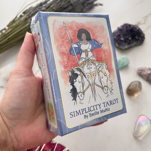 May include: A tarot card deck titled "Simplicity Tarot" by Emille Mu&ntilde;iz. The box is light blue with gold trim and features an illustration of a woman riding a chariot pulled by two horses. The background is red with white swirls.