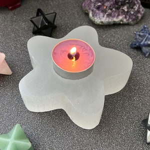 May include: A white selenite star-shaped candle holder with a pink tea light candle burning inside. The candle holder is on a dark gray surface.