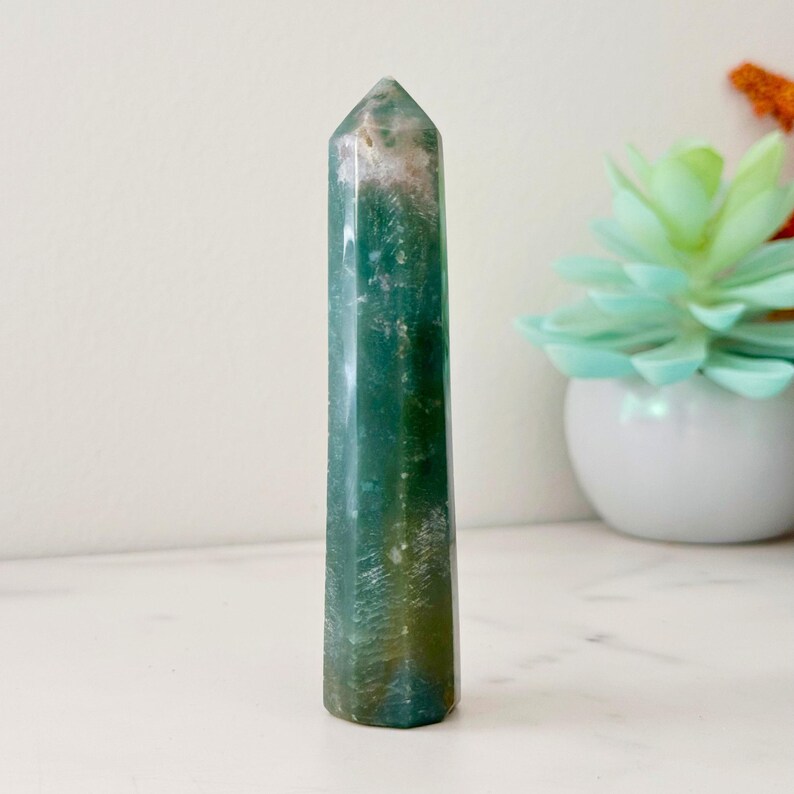 May include: A tall, polished green gemstone obelisk with white and brown inclusions. The crystal has a faceted, pointed top and a smooth, cylindrical body. The obelisk is displayed on a white surface, with a succulent plant in the background.