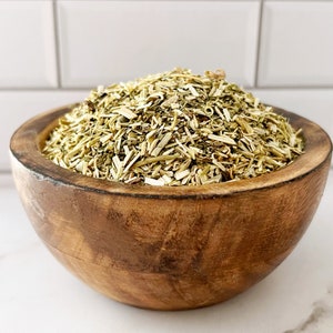 May include: A wooden bowl filled with dried, light green and brown herbs. The herbs are chopped into small pieces and look like they could be used for tea.