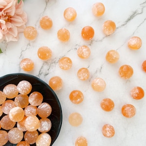 May include: A collection of orange carnelian tumbled stones. The stones are smooth and polished, with a natural, cloudy appearance. Some of the stones are arranged in a black bowl, while others are scattered on a white marble surface.