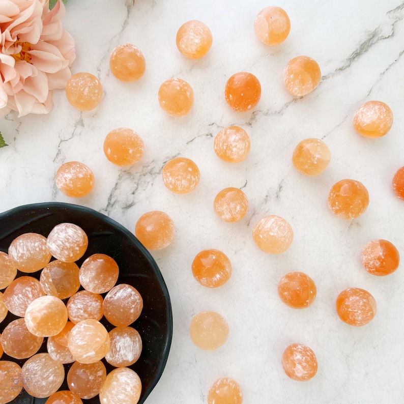 May include: A collection of orange carnelian tumbled stones. The stones are smooth and polished, with a natural, cloudy appearance. Some of the stones are arranged in a black bowl, while others are scattered on a white marble surface.