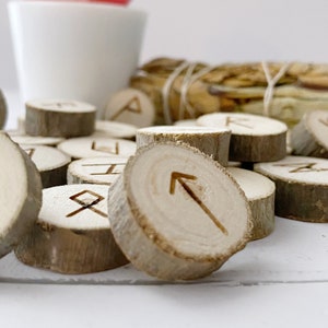 May include: A set of wooden runes with various symbols carved into them. The runes are arranged in a pile on a white surface.