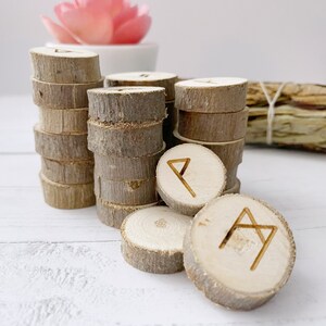 May include: A set of wooden runes with different symbols carved into them. The runes are stacked in a pile and are made of natural wood.
