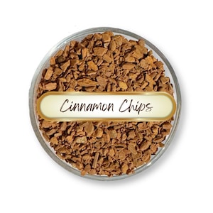 May include: A clear glass bowl filled with brown cinnamon chips. A white and gold label across the bowl reads "Cinnamon Chips" in a cursive font. The chips are small, irregularly shaped pieces.
