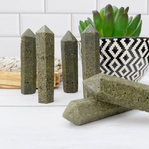 May include: Five green crystal points, also known as wands, are arranged on a white surface. Two more green crystal points are lying on their side in the foreground. The crystals are a dark green color and have a rough, textured surface.