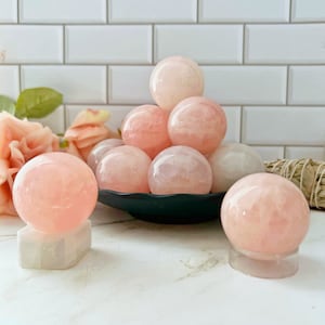 May include: A collection of pink rose quartz spheres, some on a black plate and others on white stands. The spheres are smooth and polished, with a soft pink hue.