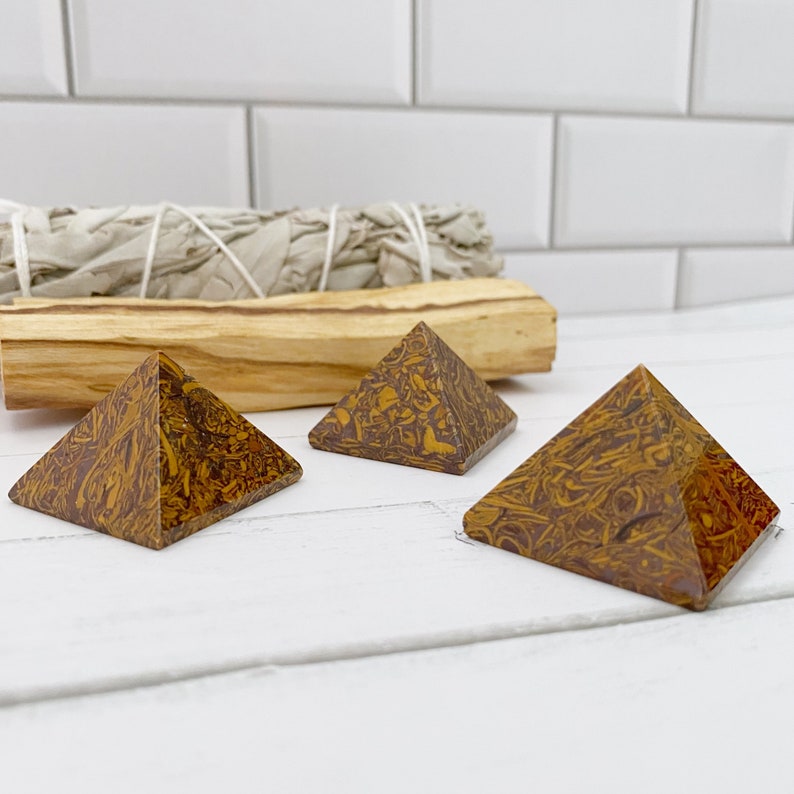 May include: Three decorative pyramid-shaped stones with a brown and yellow patterned design. The pyramids are displayed on a white surface, with a bundle of sage and a piece of palo santo wood in the background.