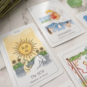 May include: Tarot cards featuring watercolor illustrations. One card shows a smiling sun, a child, and a white horse, labeled "The Sun." Another card depicts an angel and figures, labeled "Judgment." The cards are on a white surface.