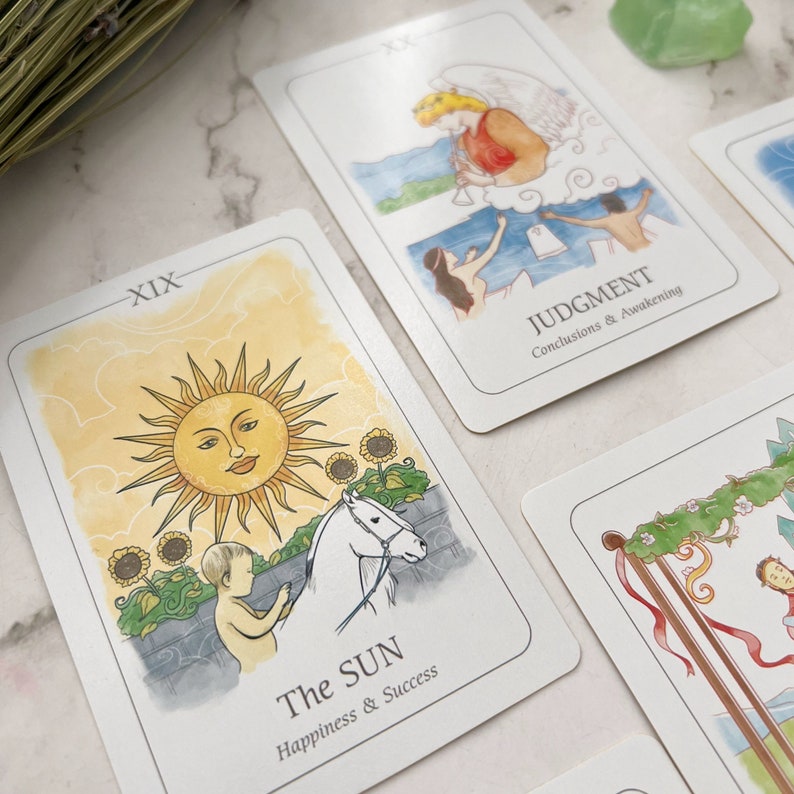 May include: Tarot cards featuring watercolor illustrations. One card shows a smiling sun, a child, and a white horse, labeled "The Sun." Another card depicts an angel and figures, labeled "Judgment." The cards are on a white surface.