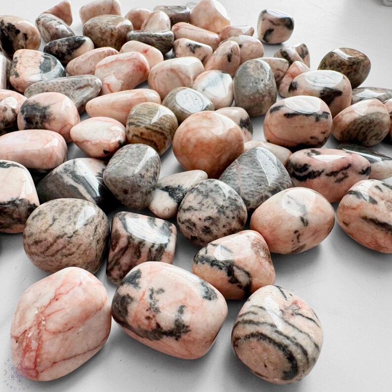 May include: A collection of polished stones in various shapes and sizes. The stones display a mix of colors, including pink, gray, and brown, with unique veining patterns. The smooth, rounded stones are scattered across a surface.