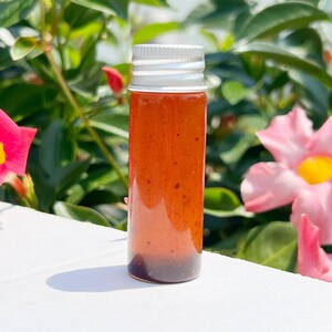 May include: A small, clear glass bottle with a silver cap, filled with a reddish-brown liquid. The bottle is standing upright against a backdrop of green foliage and pink flowers. The liquid appears to have a slightly thick consistency.