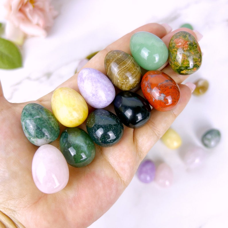 Puede incluir: Una colecci&oacute;n de huevos de piedras preciosas pulidas en varios colores, incluyendo verde, rojo, morado, amarillo y marr&oacute;n. Los huevos est&aacute;n dispuestos en una mano, con algunos huevos adicionales esparcidos sobre una superficie blanca.
