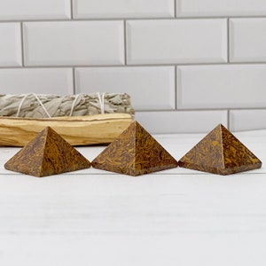 May include: Three brown and yellow patterned stone pyramids are displayed on a white surface. The pyramids are arranged in a row, each with a distinct pattern. A bundle of sage and a wooden stick are in the background.