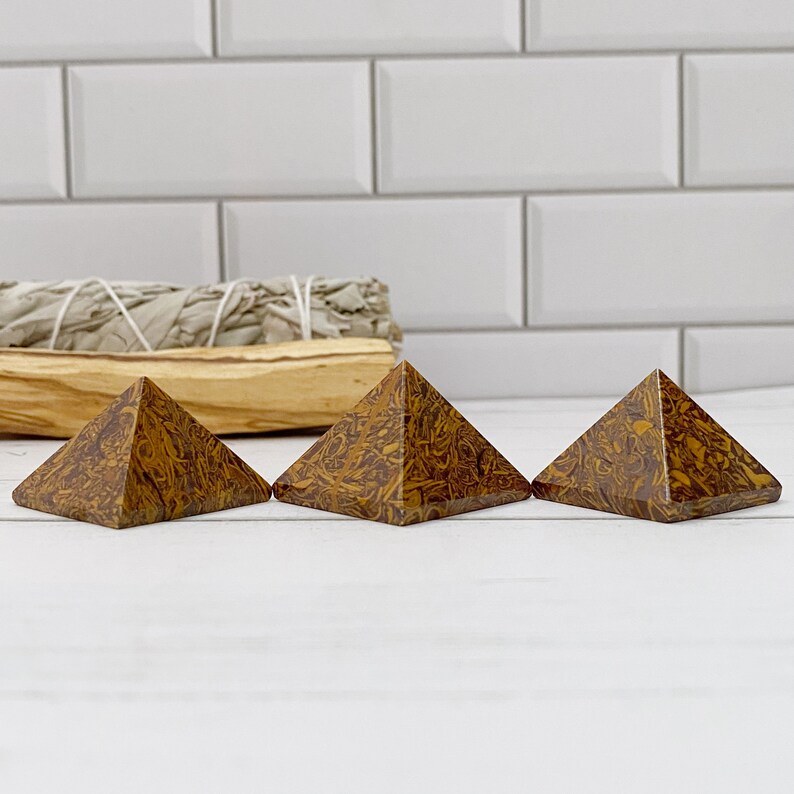 May include: Three brown and yellow patterned stone pyramids are displayed on a white surface. The pyramids are arranged in a row, each with a distinct pattern. A bundle of sage and a wooden stick are in the background.