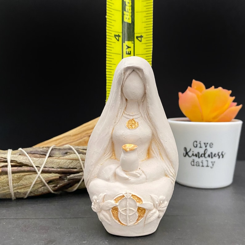 Puede incluir: Figura de color crema de una mujer sentada con un velo. La estatua mide aproximadamente 10 cm de altura y presenta detalles dorados en el collar, el pecho y la base. Una regla amarilla se coloca detr&aacute;s de la estatua.