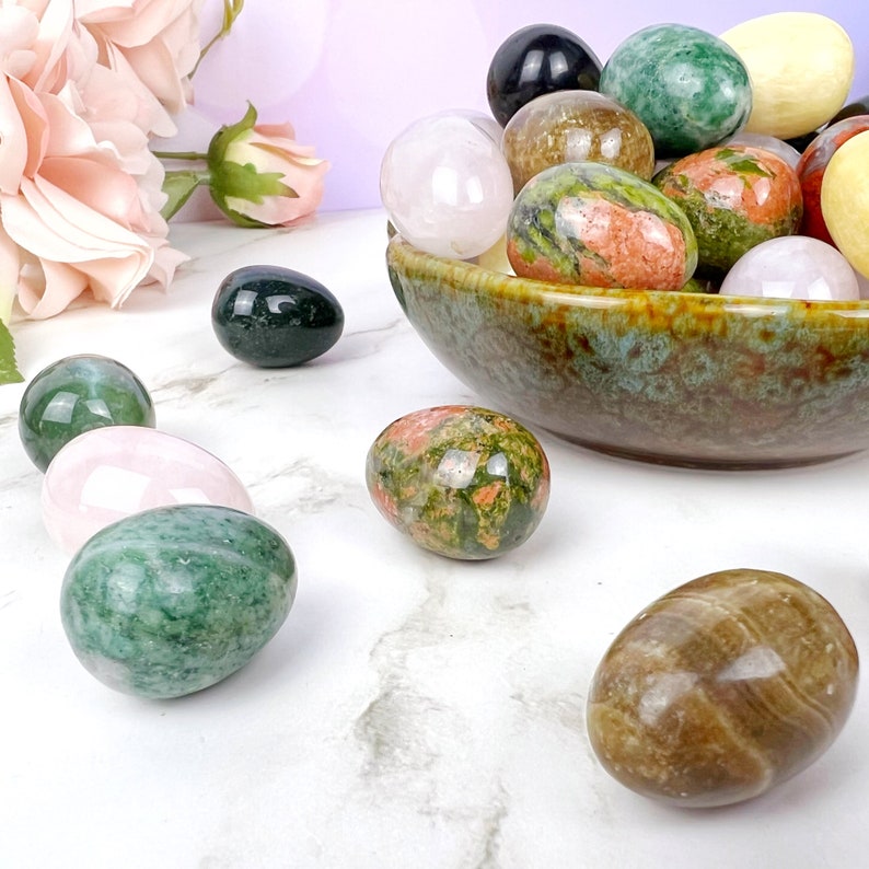 Puede incluir: Una colecci&oacute;n de huevos de piedras preciosas pulidas en varios colores, incluyendo verde, rosa, marr&oacute;n y multicolor, exhibidos en un cuenco y sobre una superficie de m&aacute;rmol blanco.
