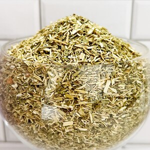 May include: A glass bowl filled with dried, light green and brown herbs. The herbs are finely chopped and appear to be a blend of different types.