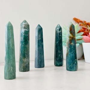 May include: Five polished gemstone obelisks in shades of green and teal. The obelisks are standing upright, with a smooth, tapered shape. The gemstones have natural patterns and variations in color. The background includes a white surface and a vase of flowers.