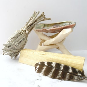 Puede incluir: Un manojo de salvia blanca, un palo santo, una pluma y una concha de mar sobre un soporte de madera.