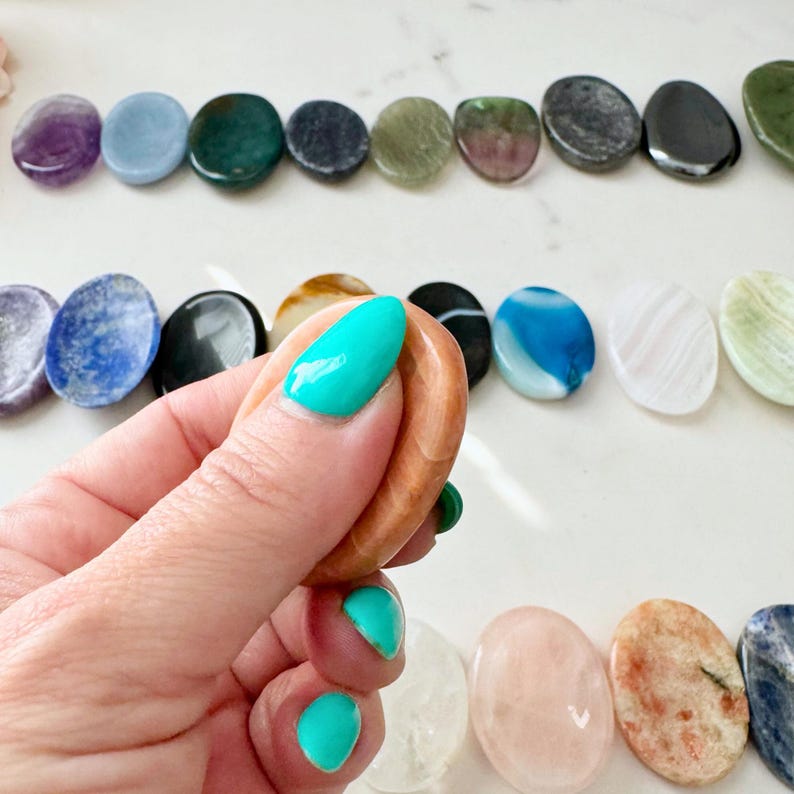 May include: A hand holding an oval-shaped, peach-colored gemstone, with a row of various polished gemstones in the background. The gemstones are in shades of purple, blue, green, black, and white. The hand has turquoise nail polish.