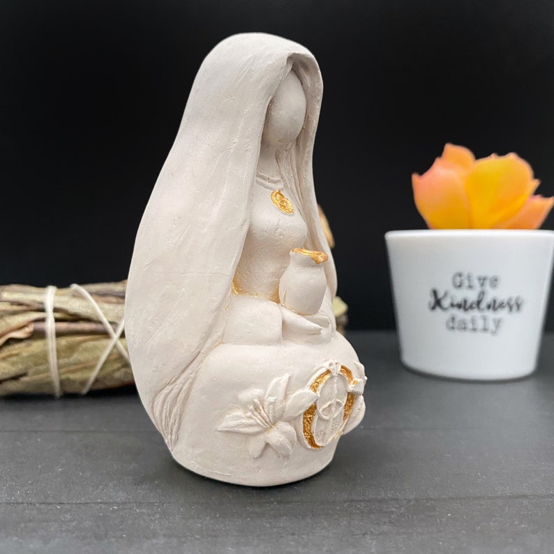 Puede incluir: Figura de color crema de una mujer sentada con cabello largo y suelto y un velo. La figura sostiene una peque&ntilde;a jarra y est&aacute; adornada con detalles dorados en el pecho y la falda. Una maceta blanca con el texto "Give Kindness daily" est&aacute; en el fondo.